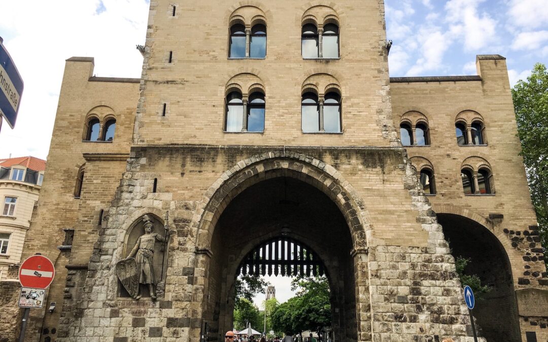 Ein großes historisches Stadttor aus Stein mit drei Torbögen und Bogenfenstern, auf dessen linker Seite die Statue einer Person in Rüstung steht, markiert den Beginn vieler Kölner Stadtführungen. Menschen gehen und radeln hindurch, im Hintergrund sind Bäume und Gebäude zu sehen.