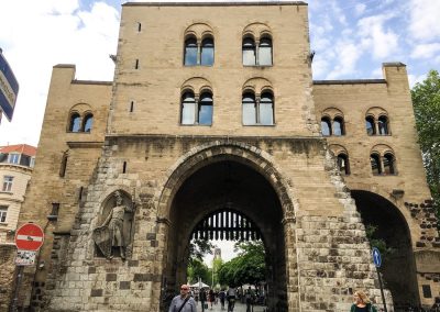 Ein großes historisches Stadttor aus Stein mit drei Torbögen und Bogenfenstern, auf dessen linker Seite die Statue einer Person in Rüstung steht, markiert den Beginn vieler Kölner Stadtführungen. Menschen gehen und radeln hindurch, im Hintergrund sind Bäume und Gebäude zu sehen.